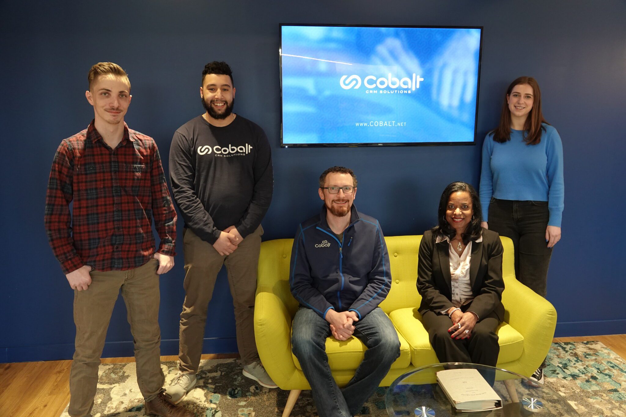 The image is a group photo of the Dynamics 365 Managed Support Services team in the lobby of Cobalt's Washington, D.C. office.